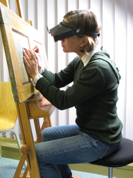 Frau Tr&ouml;ger, welche eine Lupenbrille um den Kopf gebunden hat, beim Restaurieren eines Bildes