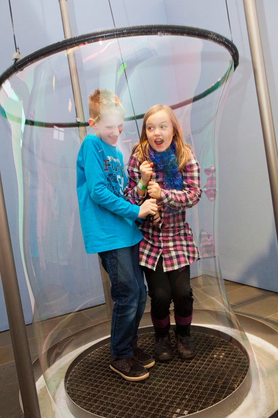 Zwei Kinder stehen auf einer Platte. Um sie herum wird ein "Seifenblasen-Schlauch" an einem Ring empor gezogen.
