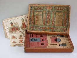 Ein h&ouml;lzener Kasten mit bunten, verschieden geformten Bausteinen. Daneben eine Bauanleitung f&uuml;r einen Turm.