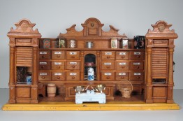 Puppenkaufladen aus Holz. Oberer Rand elegant geschwungen, in der Mitte viele Schubladen, oben auf einige Dosen. Vorne eine kleine Waage und ein Korb und ein F&auml;sschen.