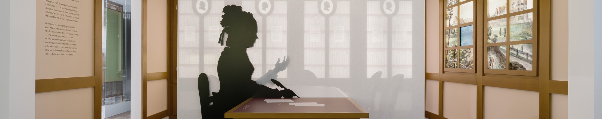 Blick in den Raum &uuml;ber Sophie von La Roche. Rechts ein Fenster angedeutet, in der Mitte ein wei&szlig;es gespanntes Tuch. Davor die Silhouette der Schriftstellerin an einem Schreibtisch mit Feder in der Hand.