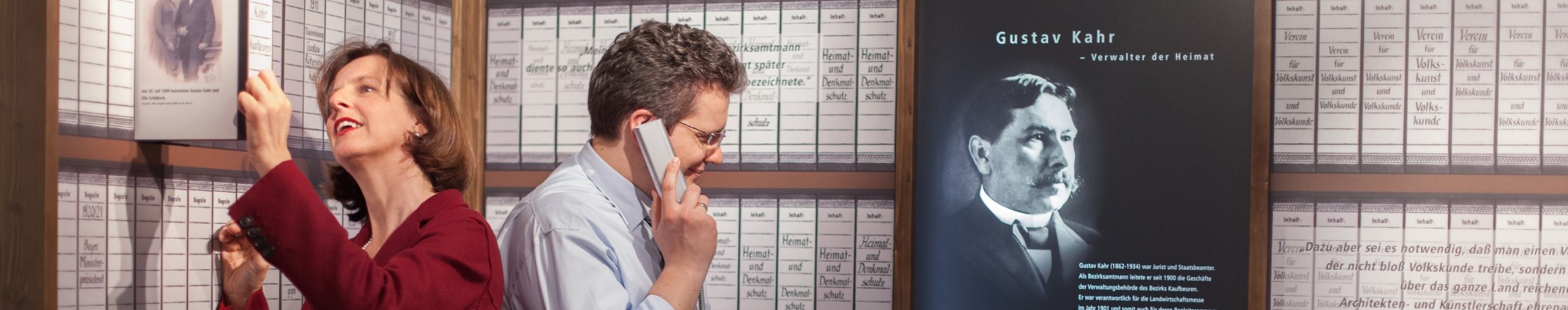 Eine Besucherin und ein Besucher sehen sich in der Abteilung &uuml;ber Gustav Kahr um: Die Frau zieht ein Buch aus der Wand, die als Buchregal gestaltet ist. Der Mann h&ouml;rt sich die Audiostation an. 