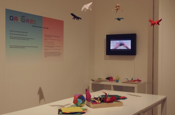 Blick in die Ausstellung: Auf einem Tisch liegen bunte Origami, in der Luft h&auml;ngen ebenfalls Origami. Im Hintergrund ist ein Bildschirm, der eine Bastelanleitung zeigt.