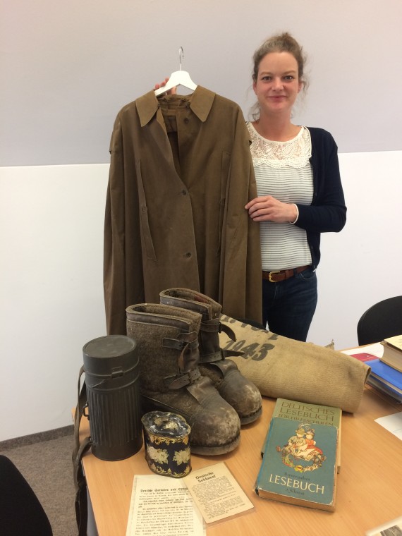 Nina Lutz h&auml;lt einen braunen BDM-Mantel in die Kamera, auf einem Tisch davor stehen Winterstiefel eines Soldaten, ein Lesebuch und weitere Objekte. 