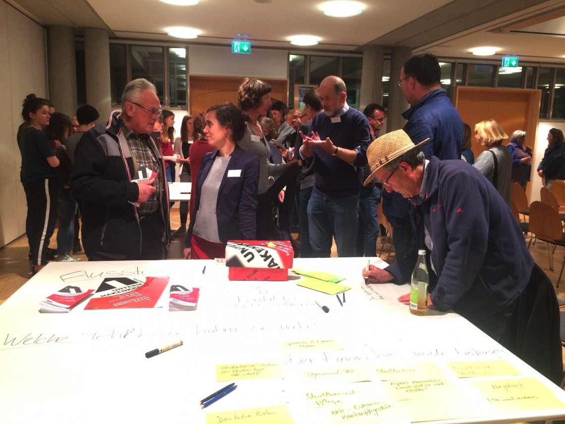 Zahlreiche Menschen im Hintergrund, im Vordergrund ein Tisch mit einer Box f&uuml;r Feedback und Anregungen. Museumsleiterin Petra Weber unterh&auml;lt sich mit einem Mann, ein anderer Mann schreibt gerade R&uuml;ckmeldung auf einen Zettel.