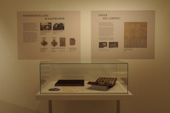 Blick in die Ausstellung: eine Glasvitrine mit Inhalt, dahinter 2 Informationstafeln &uuml;ber die Papierherstellung und "Papier aus Lumpen?"