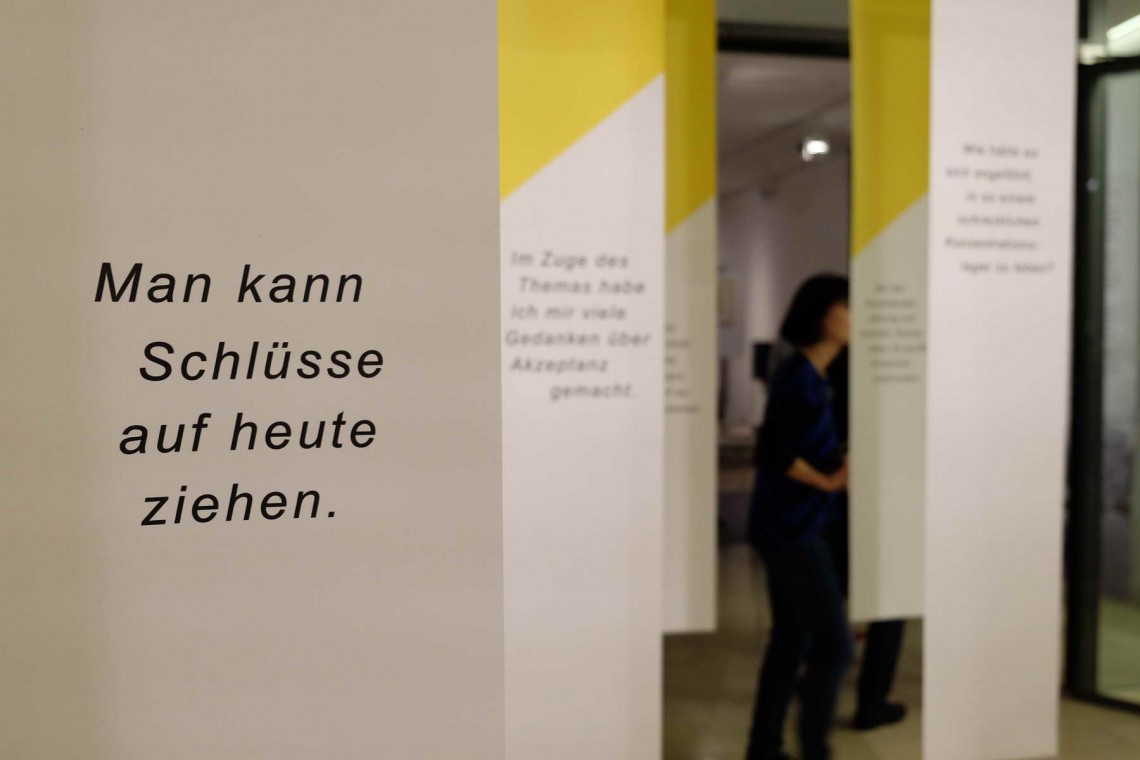 Besucher sehen sich die Themeninsel "Das Stadtmuseum" an. Auf dem Tisch liegen alte Stra&szlig;enschilder. Im Hintergrund sehen sich Menschen die Ausstellung an. Von der Decke h&auml;ngen Fahnen herab, auf denen Zitate der Besucher stehen.