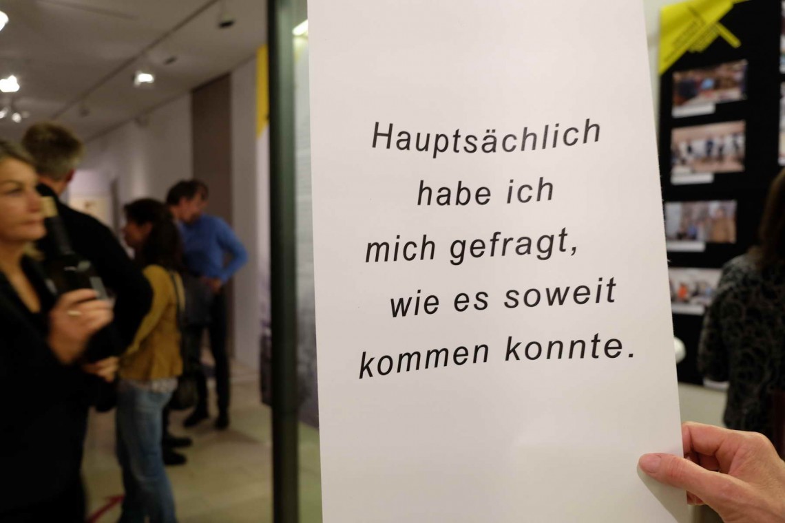 eine Hand h&auml;lt eine wei&szlig;e l&auml;ngliche Fahne in der Hand, auf der steht: "Haupts&auml;chlich habe ich mich gefragt, wie es so weit kommen konnte.". Im Hintergrund mehrere Menschen, die sich die Ausstellung ansehen 
