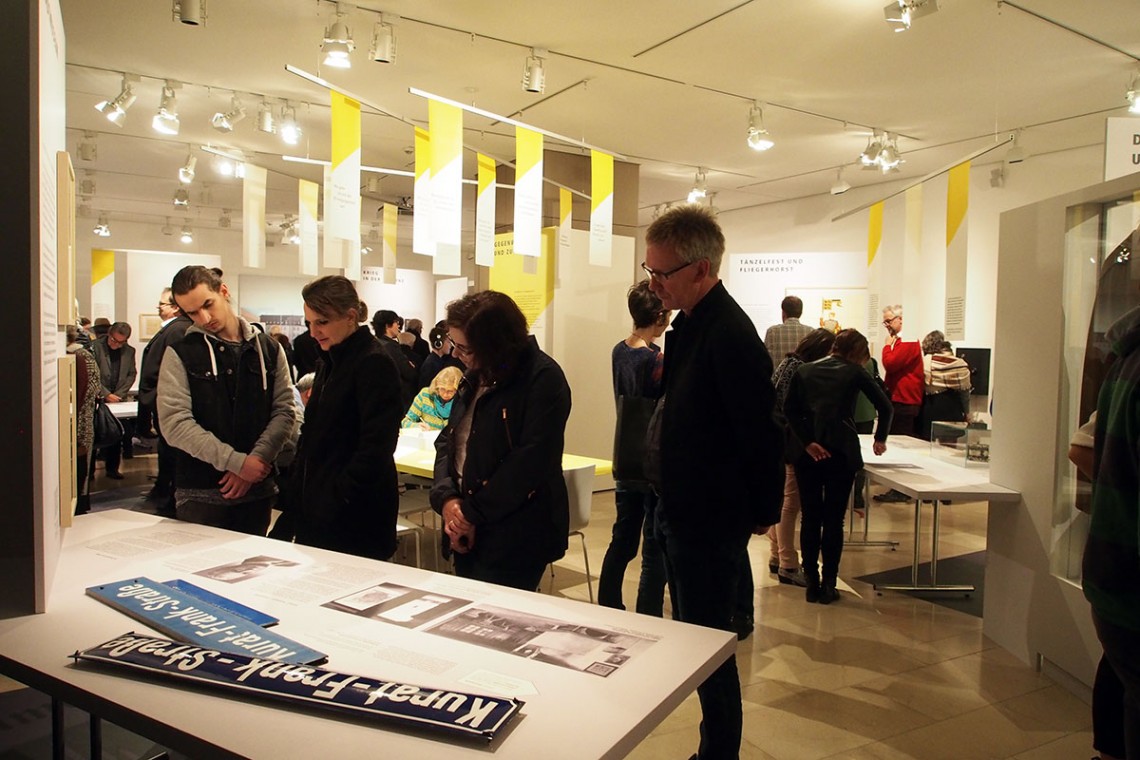 Besucher sehen sich die Themeninsel "Das Stadtmuseum" an. Auf dem Tisch liegen alte Stra&szlig;enschilder. Im Hintergrund sehen sich Menschen die Ausstellung an. Von der Decke h&auml;ngen Fahnen herab, auf denen Zitate der Besucher stehen.