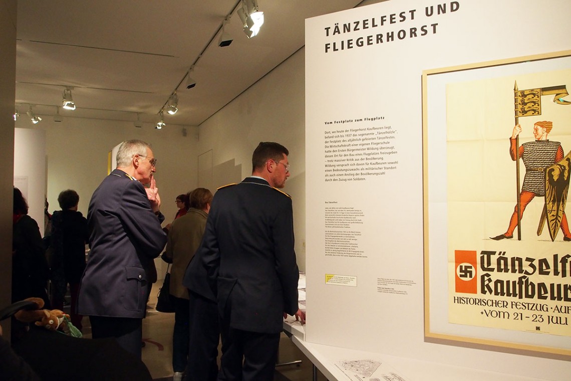 Besucher sehen sich die Themeninsel "T&auml;nzelfest und Fliegerhorst" an. Rechts ist ein Plakat zu sehen, auf dem ein Ritter in R&uuml;stung zu sehen ist, darunter ein Hakenkreuz.