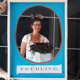 Nachstellung des Hinterglasbildes: Eine Frau mittleren Alters im Dirndl mit Blumenkranz im Haar und Blumenkorb in der Hand