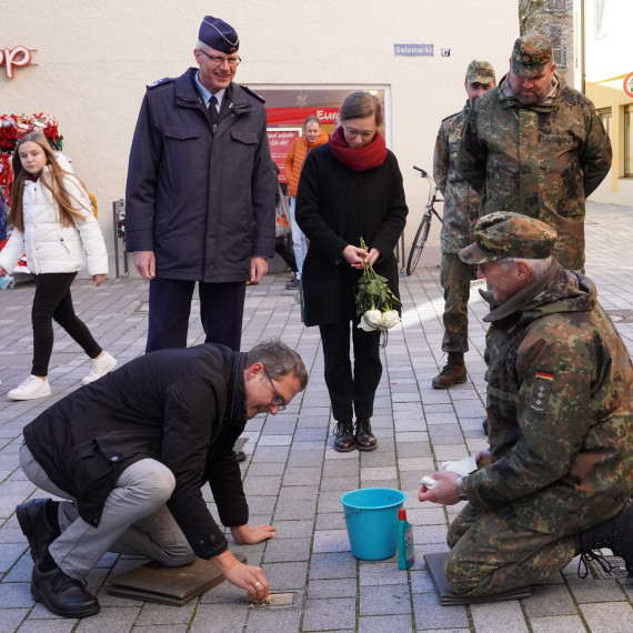 B&uuml;rgermeister Oliver Schill kniet in der Fu&szlig;g&auml;ngerzone und putzt einen Stolperstein, vor ihm ein hellblauer Eimer und eine Flasche Putzmittel. Weiters vier Vertreter der Bundeswehr und Museumsleiterin Petra Weber