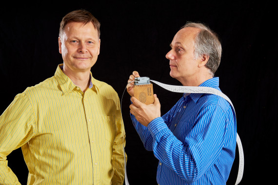 Zwei M&auml;nner, der linke mit einem gelben Hemd und der rechte mit einem blauen Hemd gekeidet. Der rechte hat ein Ger&auml;t in der Hand, aus welchem bis &uuml;ber seine rechte Schulter ein Lochstreifen kommt