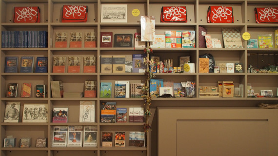 Regale des Museumsshops oben gef&uuml;llt mit roten Museumstaschen, auf der linken Seite verschiedene B&uuml;cher und auf der rechten Seite verschiedene Geschenkartikel