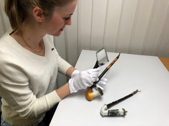 Lisa Wagner, mit wei&szlig;en Handschuhen an, inspiziert mit einer Lupe eine alte Tabakspfeife. Auf dem Tisch liegend daneben eine weitere Tabakspfeife