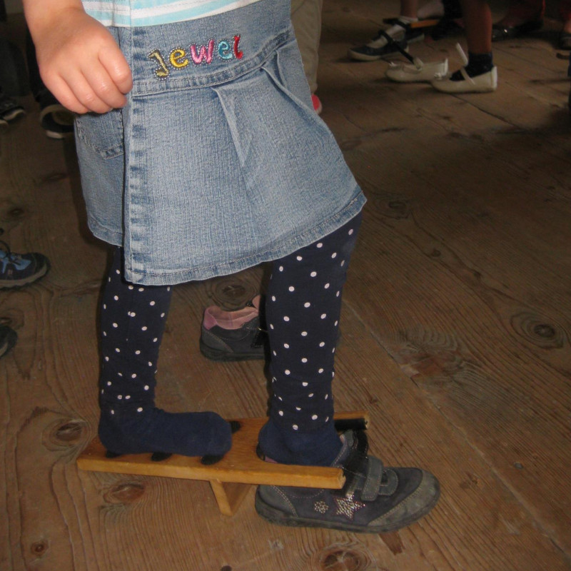 M&auml;dchen zieht sich mit einem "Stiefelknecht" den Schuh aus. Ein Stiefelknecht ist ein Holzbrett mit hufeisenf&ouml;rmigem Ausschnitt, in den man seinen Fu&szlig; steckt und so den Schuh abstreifen kann.