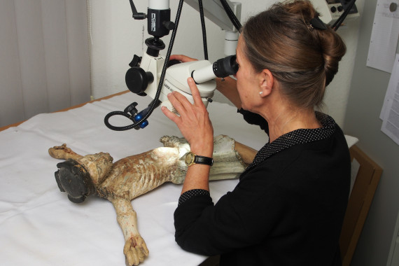 Restauratorin Heide Tr&ouml;ger schaut sich durch ein Mikroskop den vor ihr auf einem Tisch liegenden Kruzifixus an 