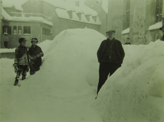 im Hintergrund H&auml;user, im Vordergrund mannshohe Schneehaufen, links stehend zwei Buben und links ein Mann, schwarz/wei&szlig;-Foto aus dem Jahr 1931