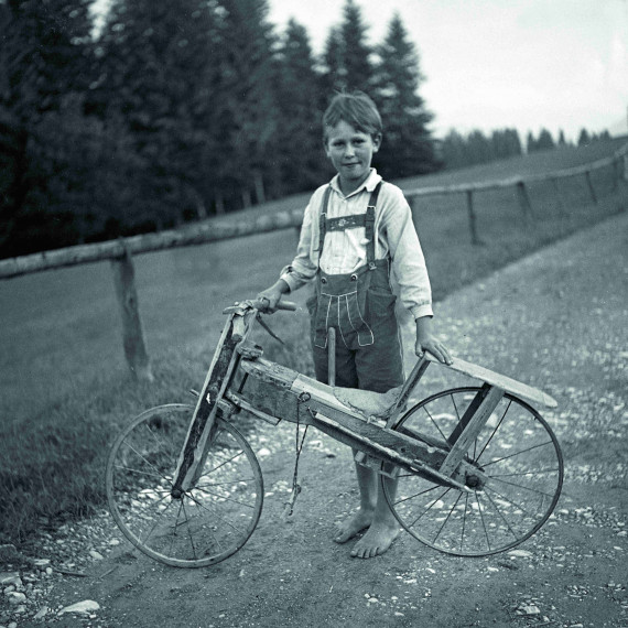 ca. f&uuml;nfj&auml;hriger Junge in Lederhose, barfu&szlig; auf einem Feldweg mit einem selbstgebautem Fahrrad