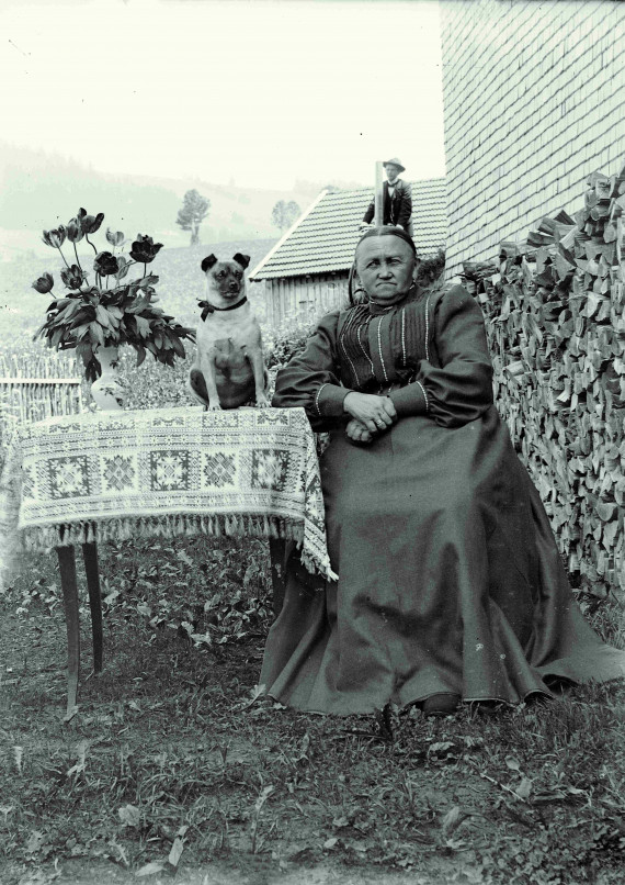 Alte Frau sitzt drau&szlig;en am Tisch, auf dem neben einer Blumenvase ein Rehpinscher sitzt. Hinter ihrem Kopf f&auml;hrt ein Radfahrer.