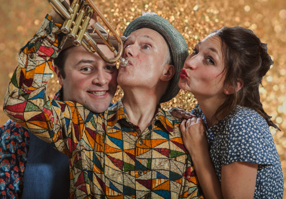 Auf der linken Seite zwei M&auml;nner, der rechte davon h&auml;lt eine Trompete an den Mund, der linke schaut zwischen Arm und Trompete hindurch, auf der rechten Seite eine Frau mit Kussmund ist aufgest&uuml;tzt auf die rechte Schulter des mittleren Mannes